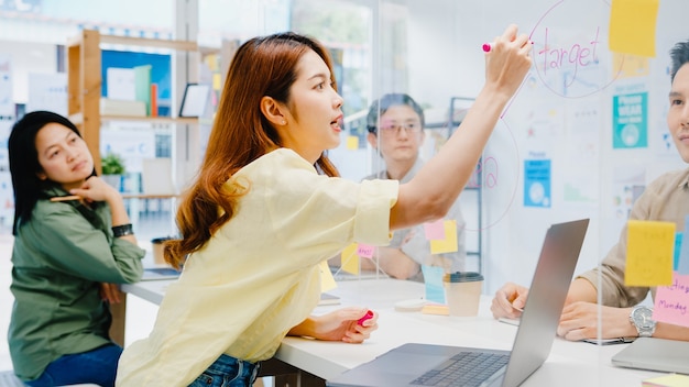 Empresários da Ásia discutindo reunião de brainstorming de negócios compartilham dados e escrevem em divisórias de acrílico no novo escritório normal. Distanciamento social do estilo de vida e trabalho após coronavírus.