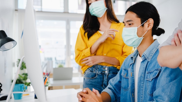 Empresários da Ásia usando apresentação de computador, reunindo-se para debater idéias sobre novos colegas de projeto e usam máscara protetora no novo escritório normal. Estilo de vida e trabalho após coronavírus.