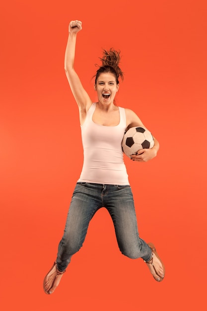 Foto grátis encaminhar para a vitória. a jovem mulher como jogador de futebol, pulando e chutando a bola no estúdio sobre um fundo vermelho. fã de futebol e o conceito do campeonato mundial. conceitos de emoções humanas