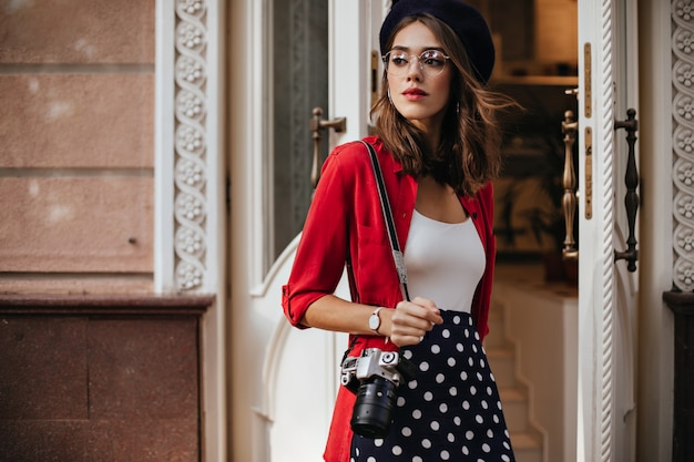 Encantador jovem fotógrafo com cabelo morena ondulado em boina, blusa branca, camisa vermelha e saia de bolinhas em pé na rua perto do café e olhando para longe