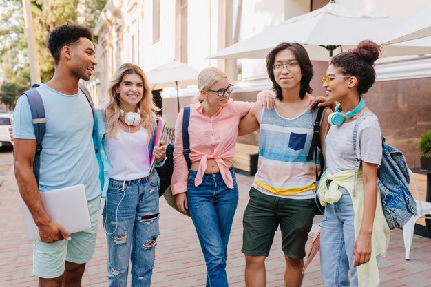 Encantadora aluna com cabelo loiro em copos em pé entre colegas de classe e olhando com sorriso para o rapaz asiático. Amigos felizes discutindo exames ao ar livre.