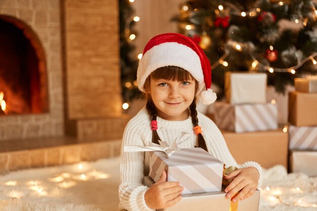 Encantadora criança feminina segurando a pilha de presentes, criança vestindo suéter branco e chapéu de Papai Noel, sentado no chão perto da árvore de Natal, caixas de presentes e lareira.