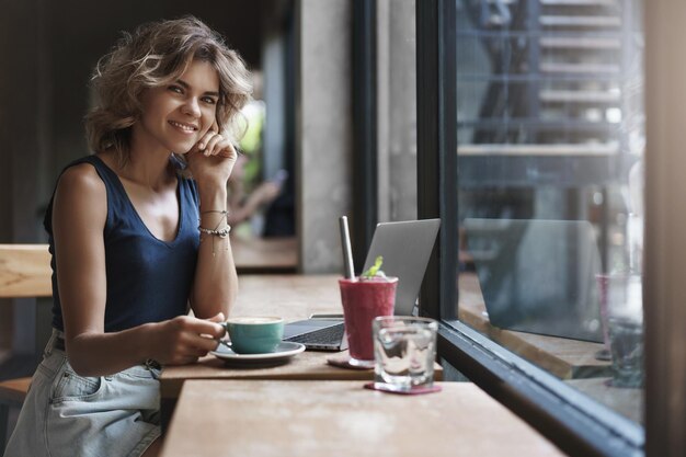 Encantadora e bem-sucedida jovem empresária que trabalha no restaurante do café sente-se perto da janela desfrute de um saboroso smoothie de café use laptop prepare o aluno do projeto estudando antes dos exames universitários