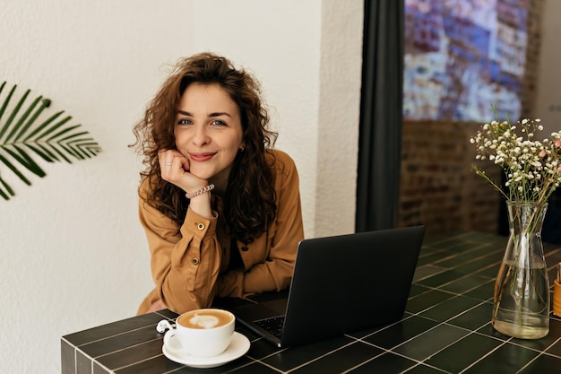 Encantadora garota adorável com cachos e batom rosa está sentada no café com café e trabalhando com laptop