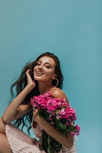 Encantadora garota de cabelos ondulados pretos com lábios grandes em brincos redondos olhando para a câmera sorrindo e segurando flores silvestres cor-de-rosa brilhantes