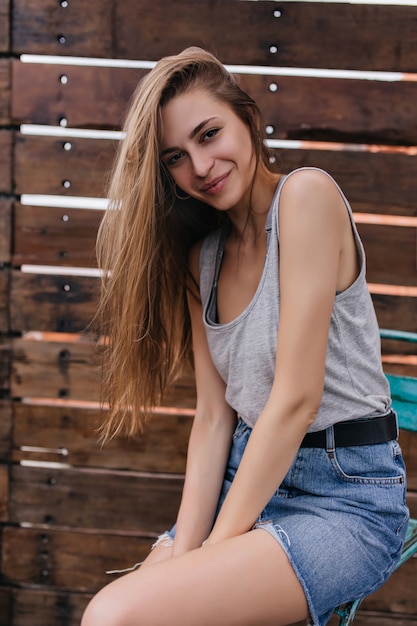 Encantadora garota europeia em saia jeans curta, posando na parede de madeira. Linda jovem com sorriso tímido, aproveitando o dia de primavera.