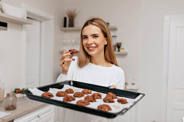 Encantadora garota sorridente com sorriso feliz segurando a assadeira com biscoitos e posando para a câmera na cozinha de casa