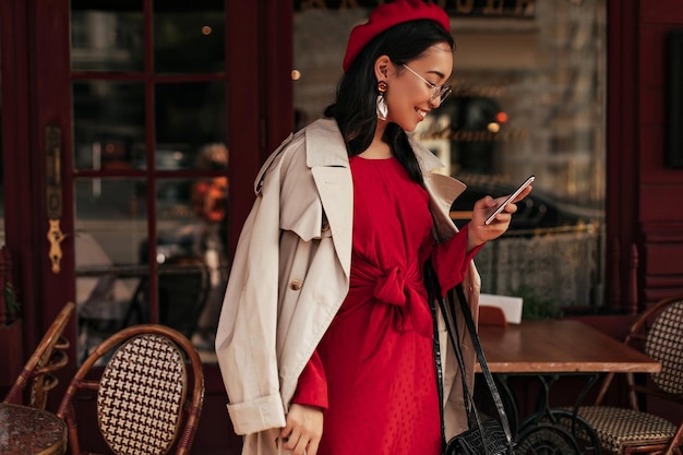 Encantadora jovem de boina de vestido vermelho e casaco bege sorrindo e enviando mensagens no celular no café de rua
