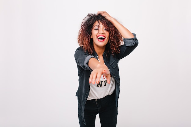 Encantadora jovem negra em jaqueta jeans, apontando com o dedo para frente. Menina encaracolada refinada com sorriso animado, posando em pé de traje da moda.