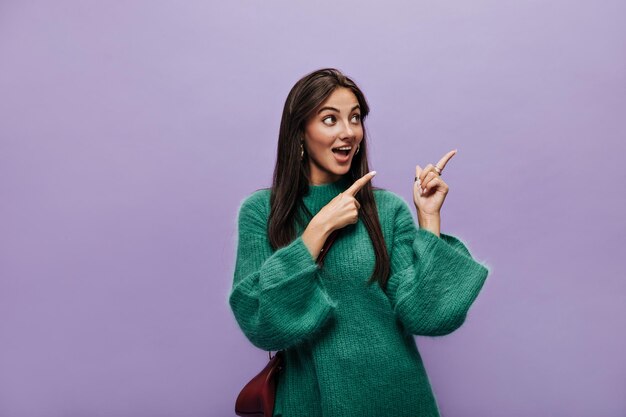 Encantadora menina morena de vestido verde aponta para o lugar para texto em fundo roxo Jovem posa em fundo isolado