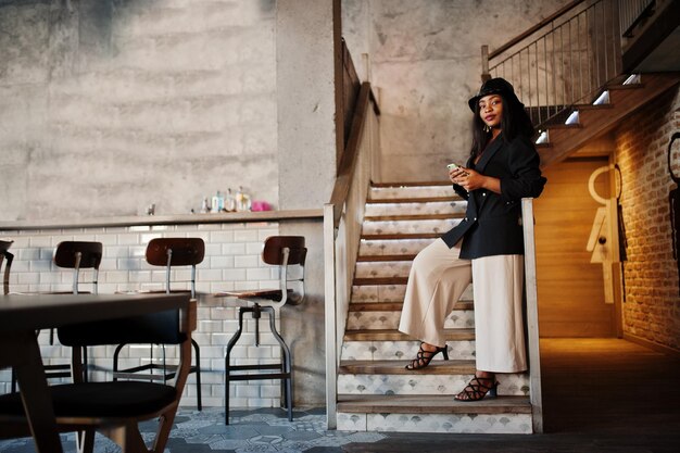 Encantadora modelo de mulher afro-americana com chapéu de jaqueta preta e bolsa de cintura relaxando no café durante o tempo livre com telefone celular