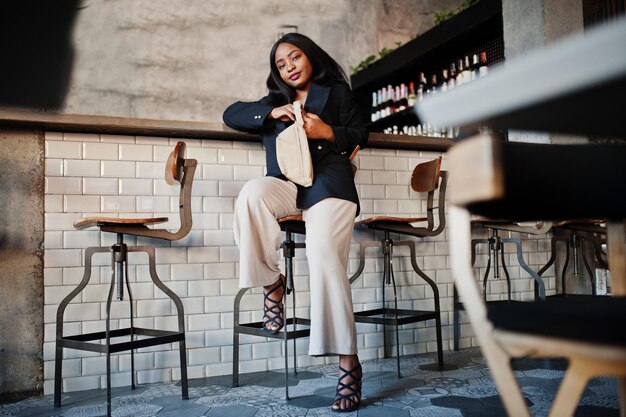 Encantadora modelo de mulher afro-americana em jaqueta preta e bolsa de cintura relaxando no café durante o tempo livre