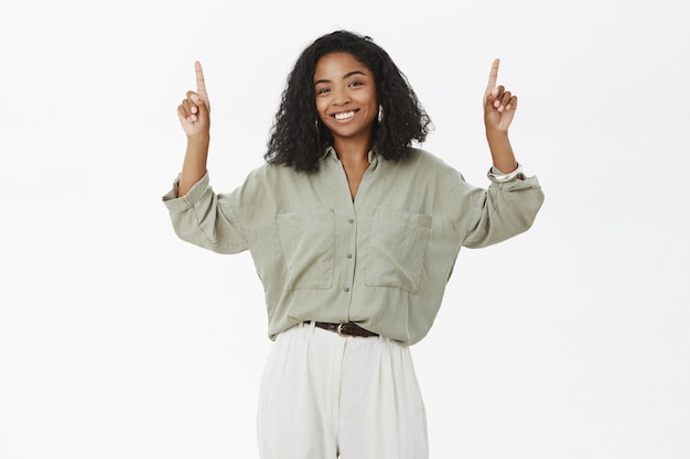 Encantadora modelo feminina de pele escura com penteado encaracolado, blusa e calça da moda levantando os braços e apontando para cima