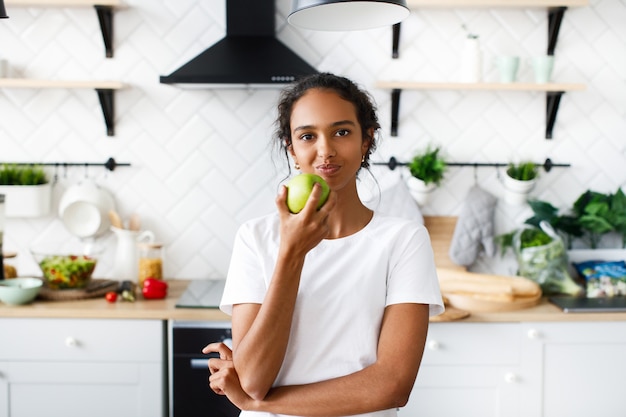 Encantadora mulher africana está comendo uma maçã verde na cozinha