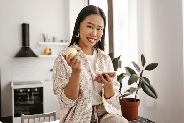 Encantadora mulher asiática de casaco de lã bege e calças olha para a câmera sorri e segura o telefone Senhora atraente com roupa elegante senta-se na mesa e segura maçã