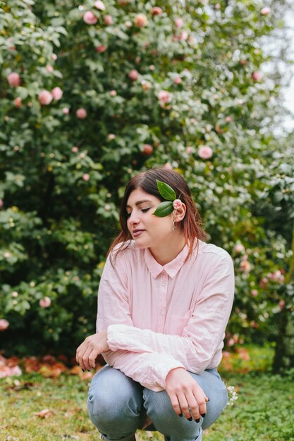 Encantadora mulher com planta no cabelo perto de flores rosa crescendo em galhos verdes