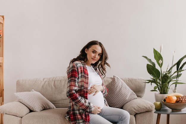 Encantadora mulher grávida olha para a câmera e toca a barriga Menina morena encaracolada em camisa xadrez e calça jeans se senta no sofá