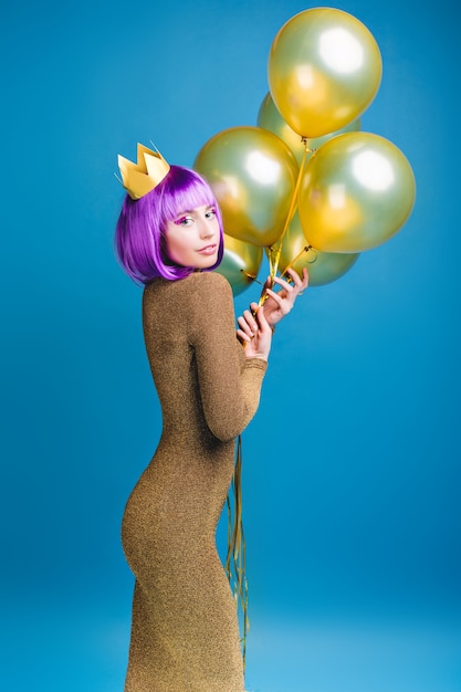 Encantadora mulher jovem e atraente na moda em vestido de luxo com balões dourados. Corte o cabelo roxo, coroa na cabeça, emoções alegres, celebração.