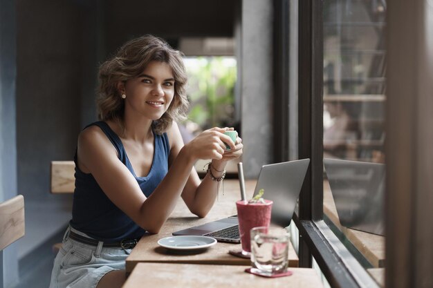 Encantadora mulher loira de cabelos encaracolados empresária sentar café bar da janela usar laptop bebendo café fazer uma pausa trabalhando freelance preparar projeto universitário conceito de nômade digital