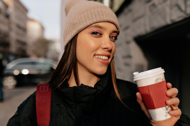 Encantadora mulher sorridente com boné bege vestido de maquiagem brilhante está segurando a xícara com café e sorrindo Muito senhora europeia relaxa no café lá fora no sol