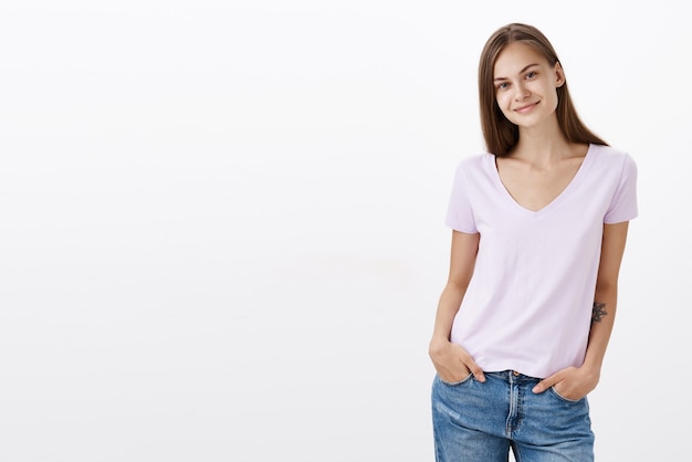 Encantadora tímida e fofa jovem morena europeia com blusa casual em pé com as mãos nos bolsos inclinando a cabeça e sorrindo com olhar amigável para conhecer novos colegas de trabalho