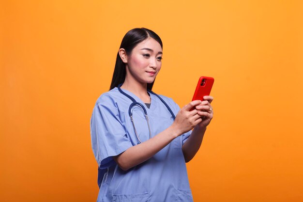 Enfermeira asiática do hospital de saúde confiante usando estetoscópio enquanto usa smartphone para enviar mensagens. Cuidador de boa aparência com dispositivo inteligente de telefone celular e instrumento médico.