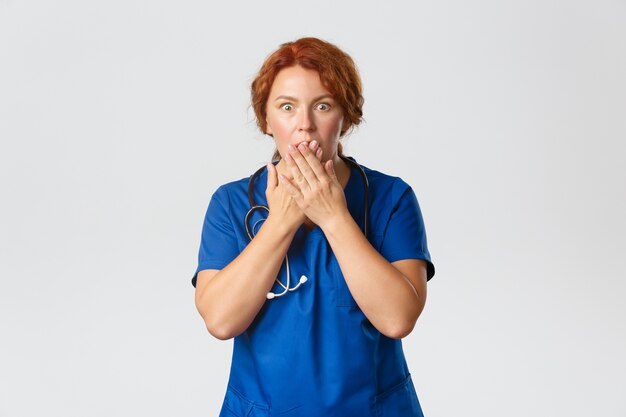 Enfermeira de meia-idade ruiva chocada e muda, médico de uniforme ouve notícias horríveis, ofegando, cobre a boca com as mãos e