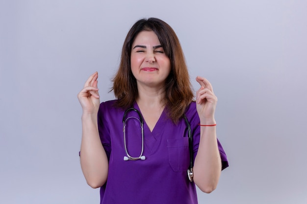 Enfermeira de meia idade vestindo uniforme e com estetoscópio com os olhos fechados levanta os dedos cruzados