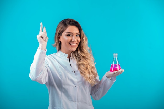 Enfermeira de uniforme branco segurando um frasco químico com líquido rosa e pede atenção.