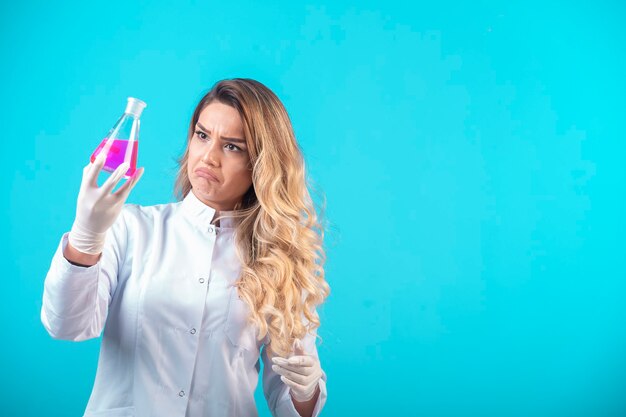 Enfermeira de uniforme branco segurando um frasco químico com um líquido rosa e parece em dúvida.