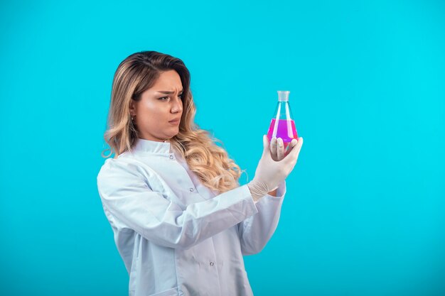 Enfermeira de uniforme branco segurando um frasco químico com um líquido rosa e tentando se lembrar.