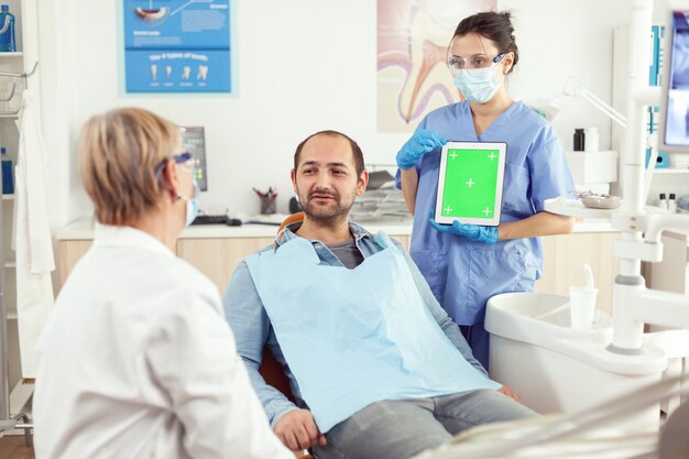 Enfermeira dentista mostrando uma tela verde para o médico sênior de estomatologia enquanto examina a dor de dente para o paciente homem sittinh na cadeira odontológica
