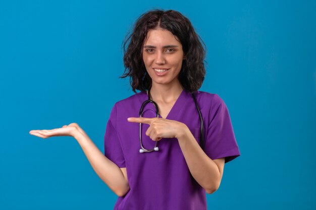 Enfermeira jovem com uniforme médico e estetoscópio apontando com o dedo para a palma da mão aberta olhando com um sorriso no rosto em pé