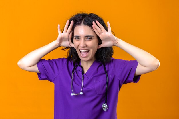 Enfermeira jovem com uniforme médico e estetoscópio parecendo animada, se divertindo, segurando as palmas das mãos abertas perto da cabeça