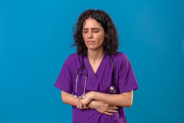 Enfermeira jovem com uniforme médico e estetoscópio parecendo doente, tocando o estômago, tendo dor em pé