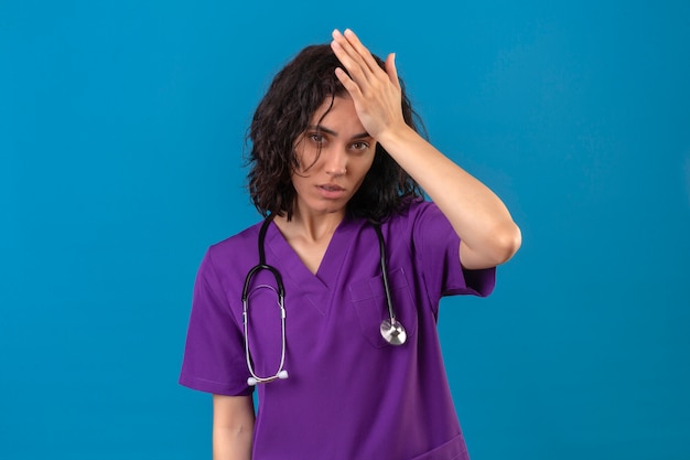 Enfermeira jovem com uniforme médico e estetoscópio parecendo insegura e com dúvida pensando com a mão na cabeça em pé
