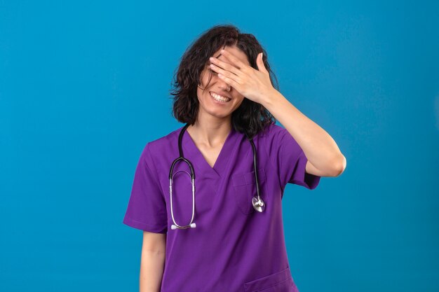 Enfermeira jovem com uniforme médico e estetoscópio sorrindo e rindo com a mão no rosto cobrindo os olhos para surpresa em pé