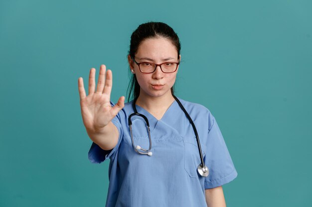 Enfermeira jovem de uniforme médico com estetoscópio no pescoço, olhando para a câmera com cara séria, fazendo o gesto de parada com a mão em pé sobre fundo azul