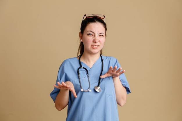 Enfermeira jovem de uniforme médico com estetoscópio no pescoço olhando para a câmera preocupada fazendo gesto de parada esticando as mãos em pé sobre fundo marrom