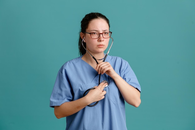 Foto grátis enfermeira jovem de uniforme médico com estetoscópio no pescoço, ouvindo seu coração batendo usando estetoscópio, parecendo confiante em pé sobre fundo azul