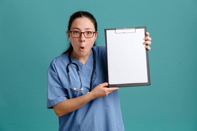 Enfermeira jovem de uniforme médico com estetoscópio no pescoço, segurando a prancheta com página em branco, olhando para a câmera espantada e surpresa em pé sobre fundo azul