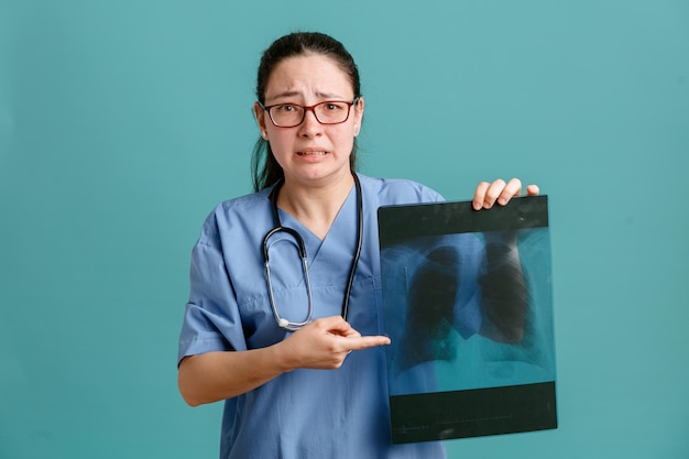 Foto grátis enfermeira jovem de uniforme médico com estetoscópio no pescoço, segurando o raio-x do pulmão apontando com o dedo indicador para ele sendo preocupado e assustado em pé sobre fundo azul