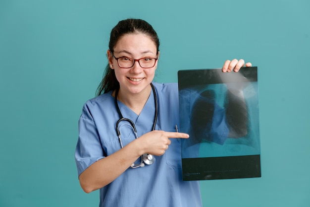 Enfermeira jovem de uniforme médico com estetoscópio no pescoço, segurando o raio-x do pulmão apontando com o dedo indicador para ele sorrindo com cara de feliz em pé sobre fundo azul