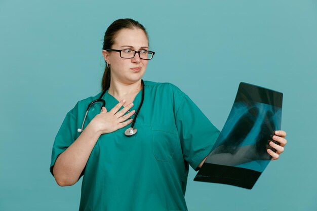 Enfermeira jovem de uniforme médico com estetoscópio no pescoço, segurando o raio-x do pulmão, olhando para ele sendo preocupado e confuso em pé sobre fundo azul