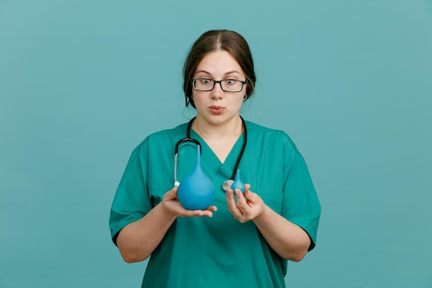 Enfermeira jovem em uniforme médico com estetoscópio no pescoço segurando enema olhando espantado e surpreso em pé sobre fundo azul