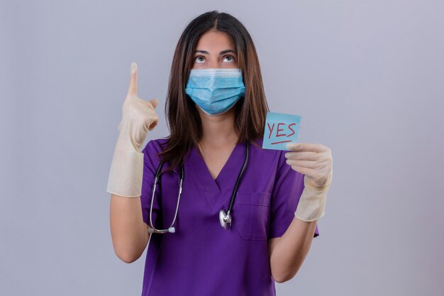 Enfermeira jovem vestindo luvas de máscara protetora de uniforme médico e com estetoscópio segurando um papel lembrete com a palavra sim apontando com o dedo para cima