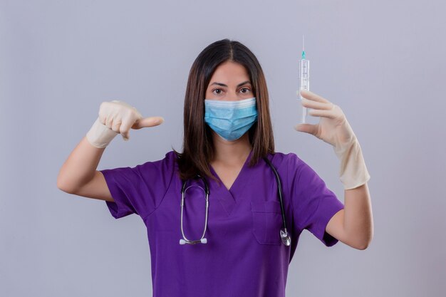 Enfermeira jovem vestindo luvas de máscara protetora de uniforme médico e com um estetoscópio segurando uma seringa apontando para ele com o polegar, olhando com expressão séria e confiante