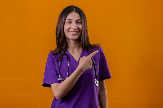 Enfermeira jovem vestindo uniforme e com estetoscópio olhando apontando para o lado com o dedo indicador com um sorriso no rosto em pé