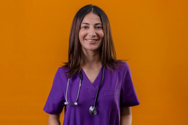 Enfermeira jovem vestindo uniforme e estetoscópio em pé com uma cara feliz e um sorriso confiante olhando