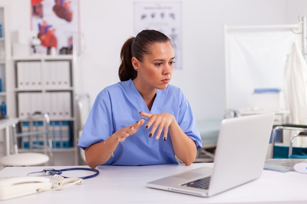 Enfermeira no consultório do hospital, lendo o tratamento do paciente no laptop. Profissional de saúde sentado na mesa usando o computador na clínica moderna, olhando para o monitor, medicina.